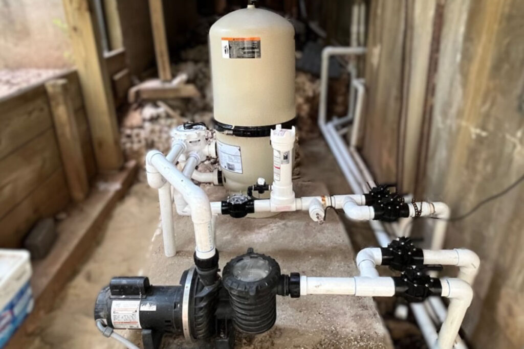 Pool filtration and circulation system with a beige filter canister, white PVC piping, and a black pool pump on a concrete base.