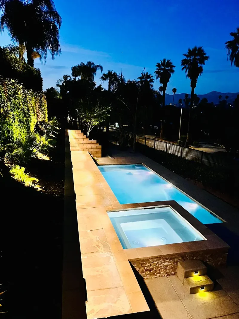 Rectangular pool with a square in-ground spa at night, illuminated stone deck and palm trees in a garden setting.
