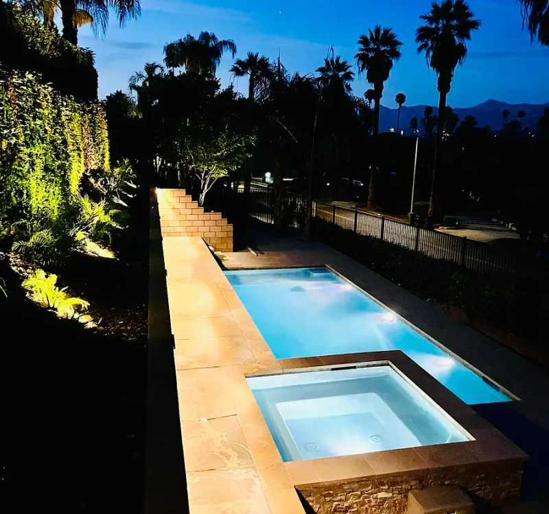 Rectangular pool with a square in-ground spa at night, illuminated stone deck and palm trees in a garden setting.